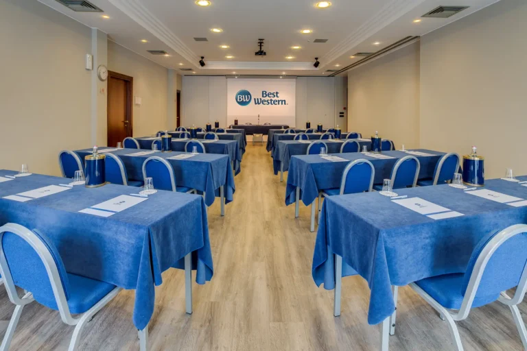 Sala meeting con disposizione a banchi di scuola, tavolo relatori sul fondo, pavimento in parquet e porta d'ingresso a sinistra