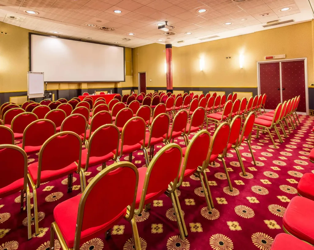 Nell'immagine si vede la Sala Meeting Diamante dell'hotel. Il pavimento della sala è di moquette rossa con decori oro. Ci sono delle file di sedie rosse rivolte verso un tavolo coperto con una tovaglia rossa. Accanto al tavolo c'è una lavagna a fogli mobili e sul muro dietro uno schermo per proiettore.