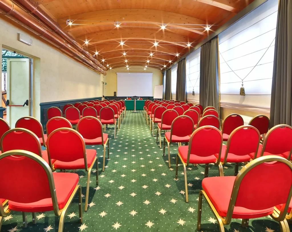 Nell'immagine si vede la Sala Meeting Rubino dell'hotel. La sala presenta un pavimento di moquette verde con decorazioni oro e un soffitto di legno a volta. Sono presenti delle file di sedie rosse rivolte verso il fondo della sala dove si trova un tavolo ricoperto con una tovaglia verde e due sedie dietro di esso. Il muro in fondo è coperto da un telo per proiezione e sul lato destro ci sono delle grandi finestre coperte con delle tende a pacchetto bianche.