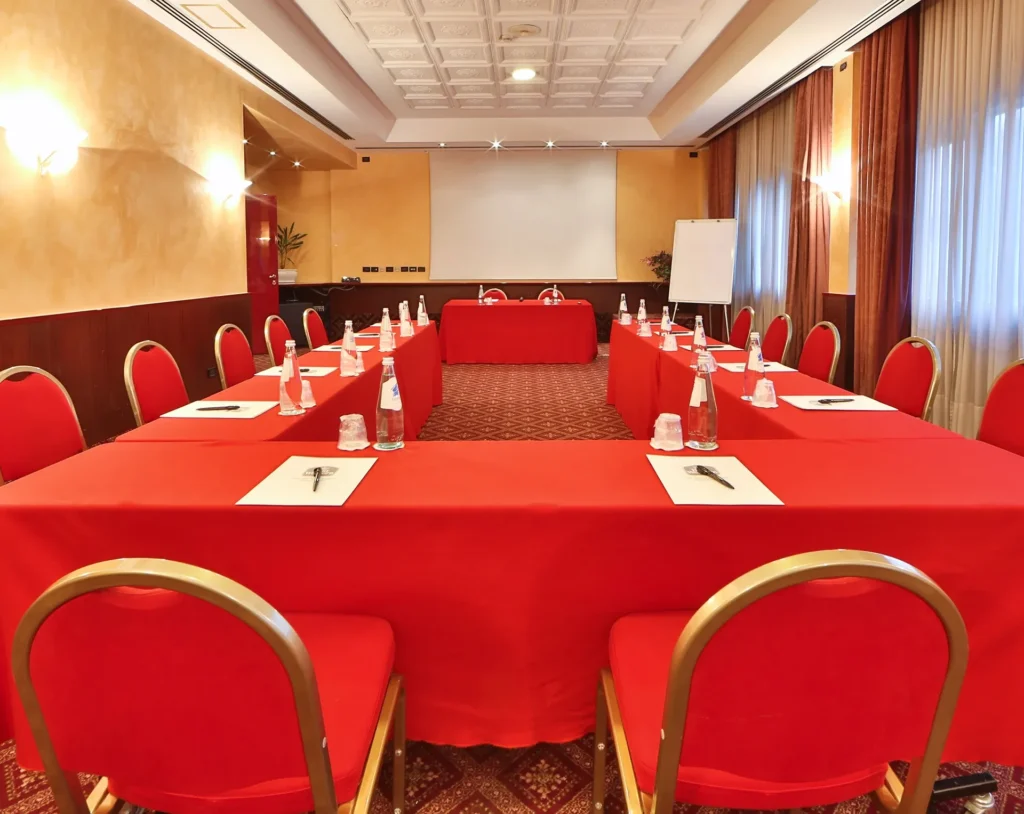 Nell'immagine si vede la Sala Meeting Corallo dell'hotel. Il pavimento è di moquette con motivi persiani rossi e oro. Si vede un tavolo a a ferro di cavallo ricoperto con una tovaglia rossa e circondato da sedie rosse. Sul tavolo sono presenti delle bottiglie, dei bicchieri di plastica e delle cartelline e penne. Sullo sfondo c'è un tavolo con due sedie. Sul muro dietro il tavolo c'è uno schermo per proiezione e accanto una lavagna a fogli mobili. Il lato destro della stanza presenta delle finestre con tende.