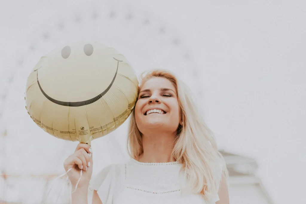 Nell'immagine una ragazza sorridente tiene nella mano un palloncino giallo con disegnato uno smile. Sullo sfondo si intravede una ruota panoramica.