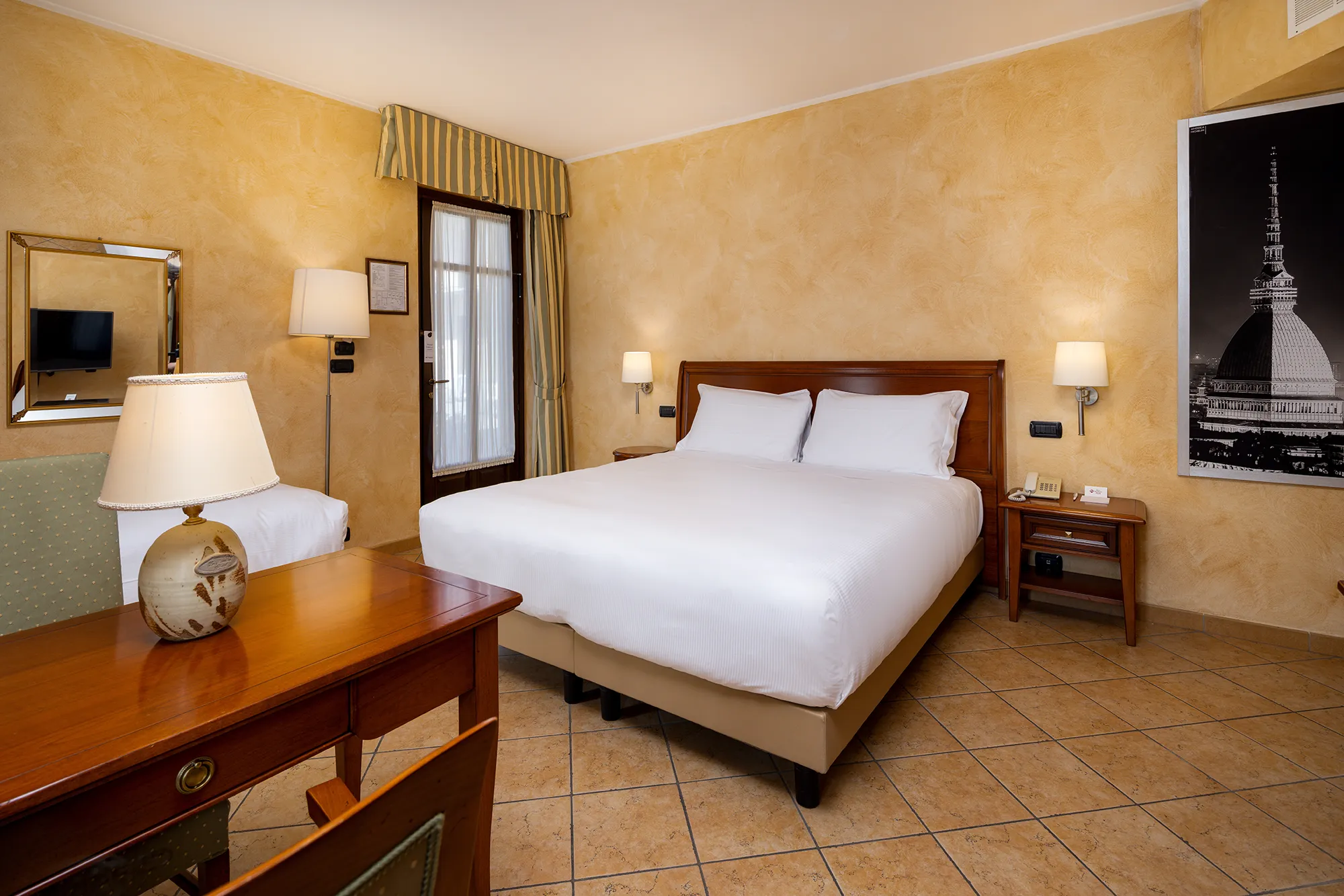 Pictured is the hotel's standard triple room. In the center of the room is the double bed with wooden nightstands and wall sconces on either side. On the wall is a post with a picture of the Mole Antonelliana. A wooden desk can be seen in the foreground with a lamp above it. Behind is a glimpse of the single bed and above it hanging on the wall is a mirror. In the corner is a French window.