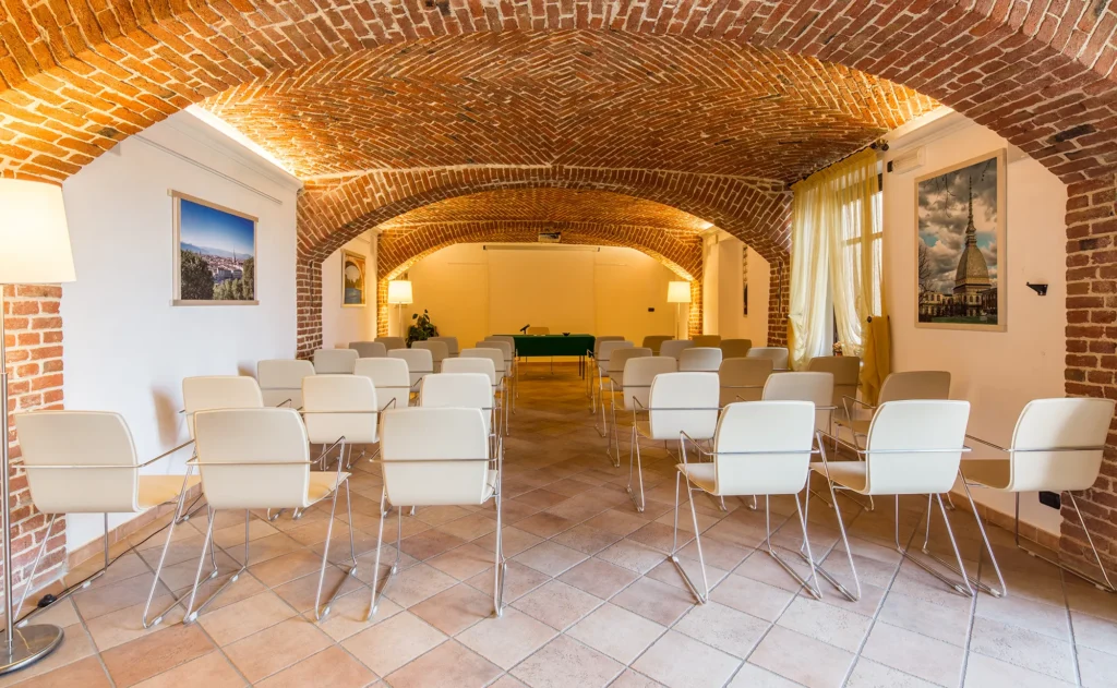 Nell'immagine la sala meeting dell'hotel con volte in mattoncini. In primo piano ci sono delle file di sedie bianche. In fondo alla sala c'è un tavolo con tovaglia verde e microfono.