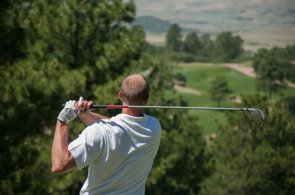 Nell'immagine si vede un uomo che ha appena terminato un tiro di golf e tiene con entrambe le mani una mazza da golf argentata. L'uomo indossa un guanto bianco e una polo bianca.