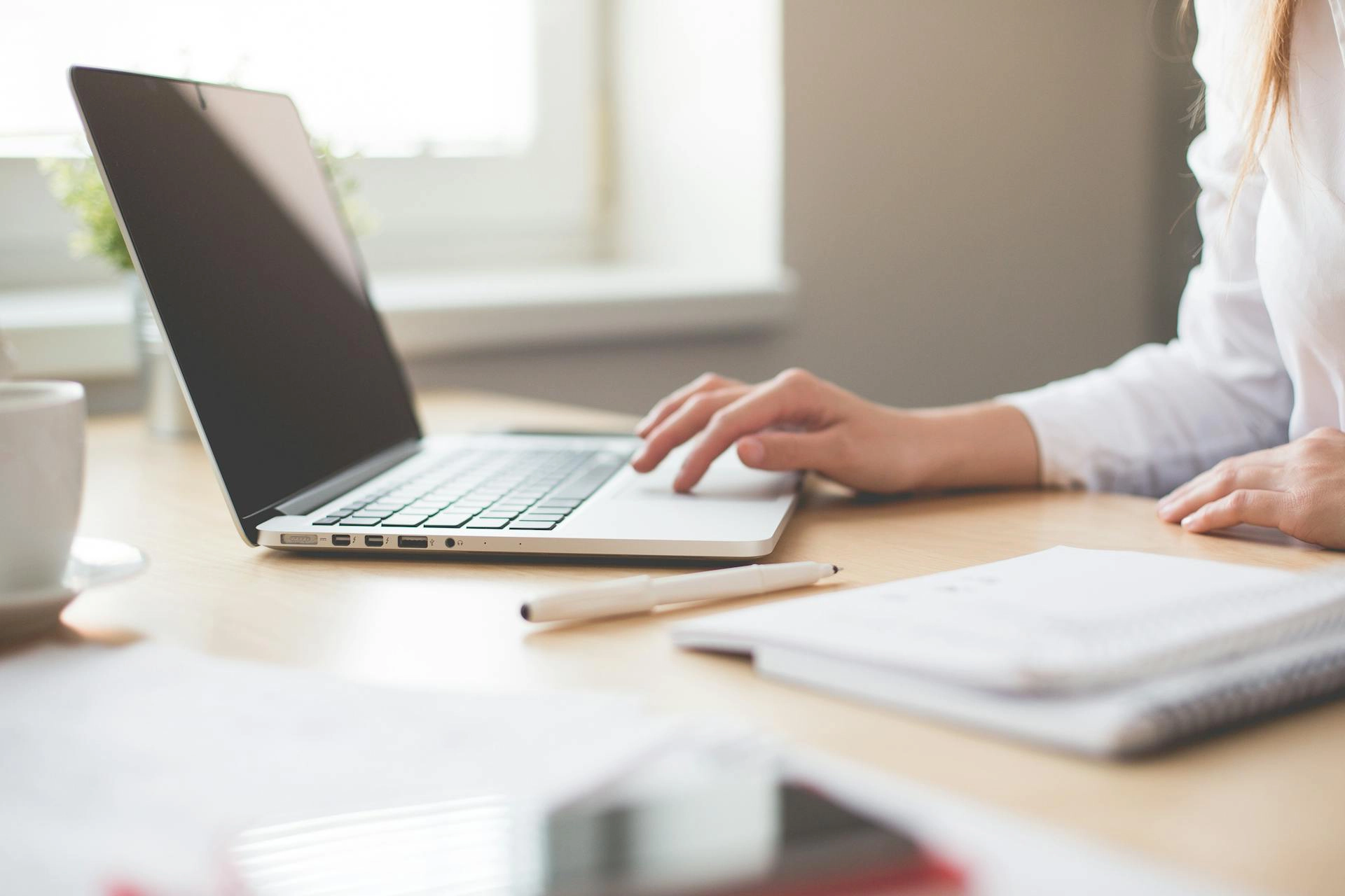 In the image you see the hand of a woman working on a laptop. Next to it can be seen a pen, a notepad and a cup.