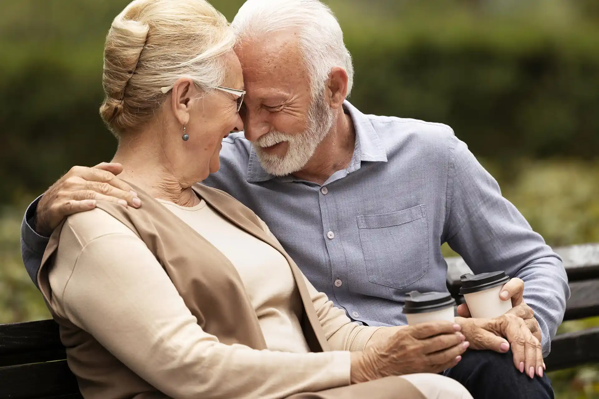 In the picture two old men sitting on a bench are smiling and resting their foreheads against each other.