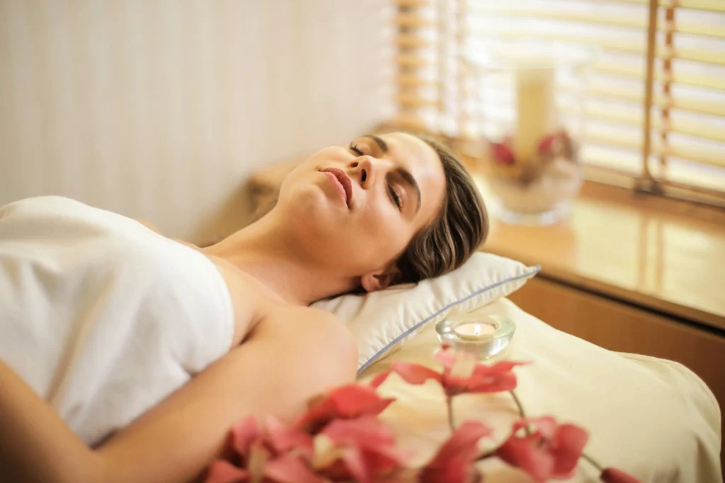 In the image, a girl is lying with her eyes closed on a massage table with a pad under her head. Next to her is a small lit candle.