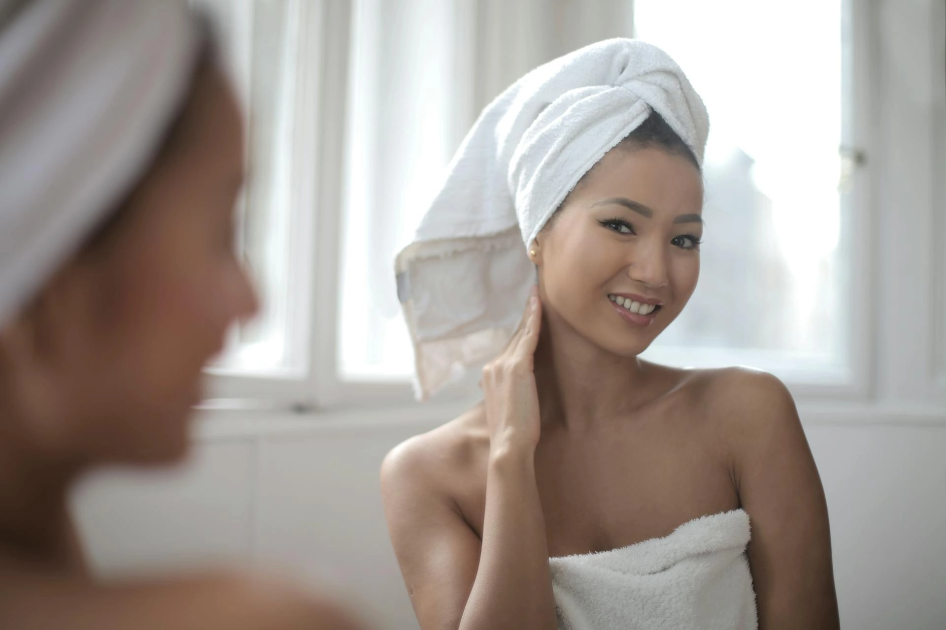 In the picture a girl looks at herself in the mirror and smiles. She wears a white towel over her hair and is covered by another towel.