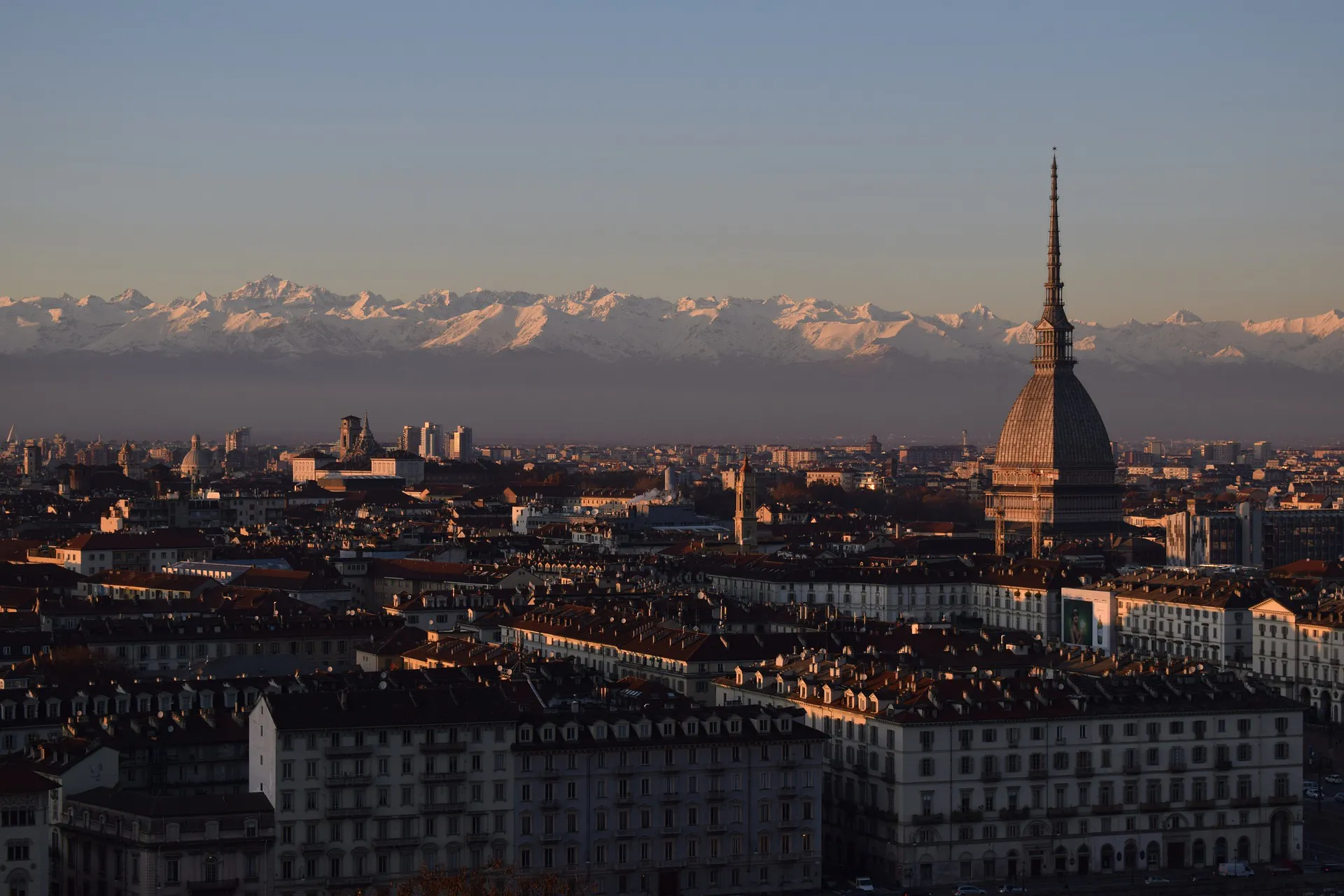 The image shows the skyline of Turin with the Mole Antonelliana in the foreground and the snow-capped Alps in the background