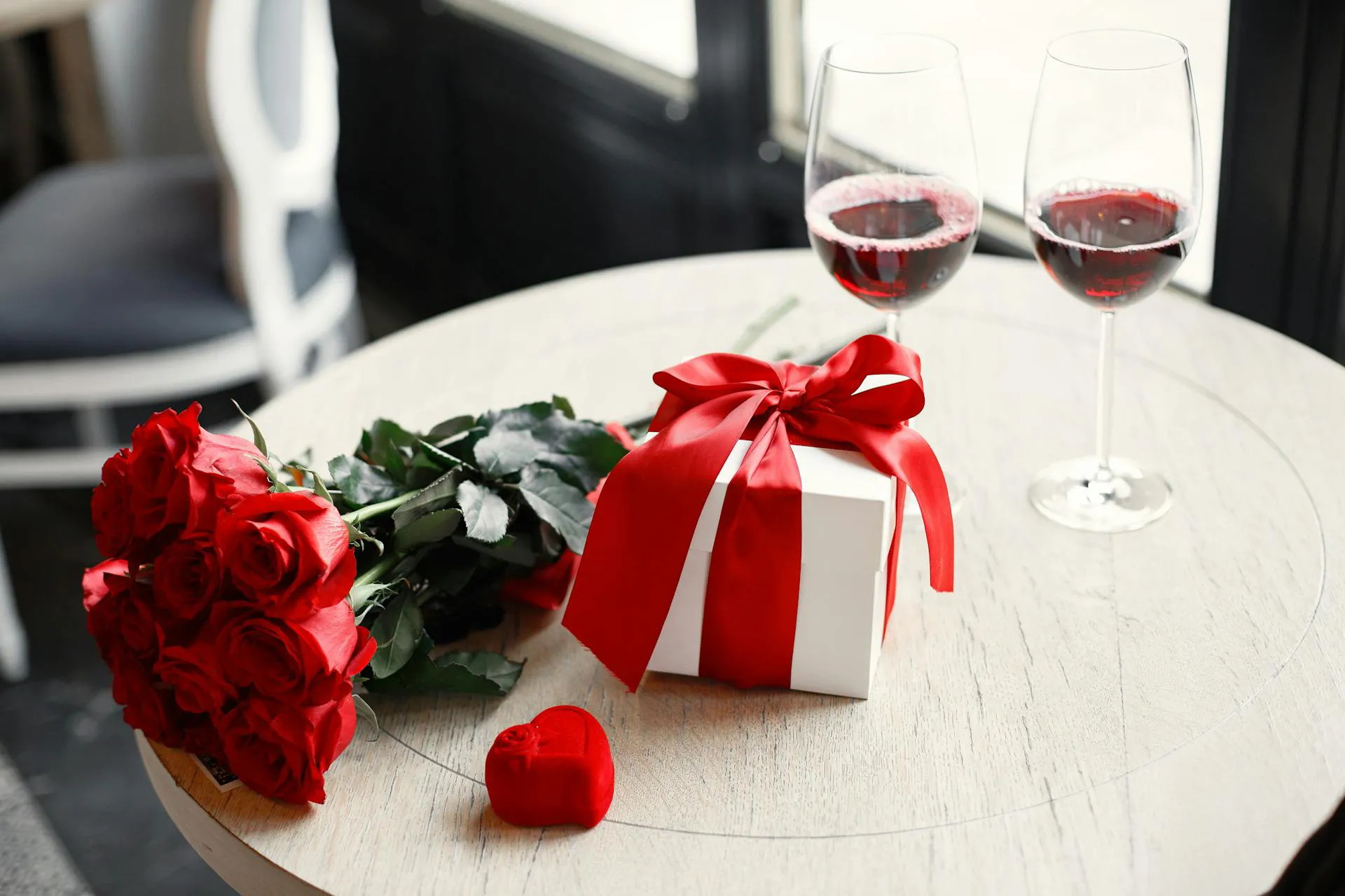 The picture shows a gift pack with a red bow, a bouquet of red roses, a red heart-shaped chocolate and, behind it, two glasses of red wine. Everything is placed on a small round wooden table.