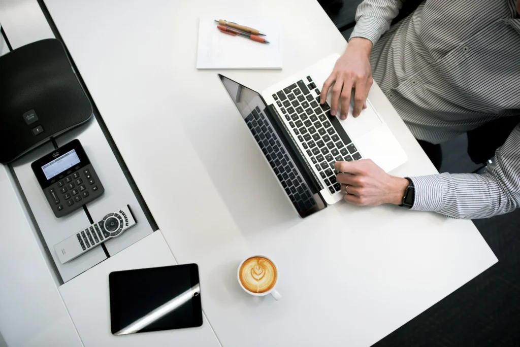 Nell'immagine un uomo con una camicia a righe è seduto a un tavolo e lavora a un pc. Accanto al pc c'è un block notes con due penne e, sulla sinistra sul tavolo, una tazza di cappuccino, un tablet nero, un telecomando, un tastierino e una cassa.